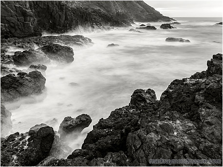 Fine art black and white long exposure photograph of waves crashing on a rugged bay