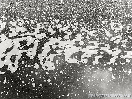 Fine art black and white photo of foam and reflections of the clouds in a roadside puddle