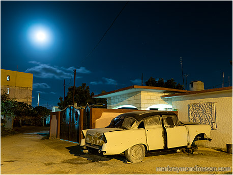 Fine art photograph showing an ancient car propped on blocks in the night with a hazy full moon in the background
