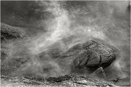 Abstract black and white photograph of misty water crashing over rough, luminescent grey rocks