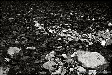 Fine art black and white photograph of pale and dark rocks in a flat, calm river