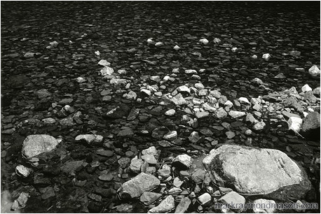 Fine art black and white photograph of pale and dark rocks in a flat, calm river