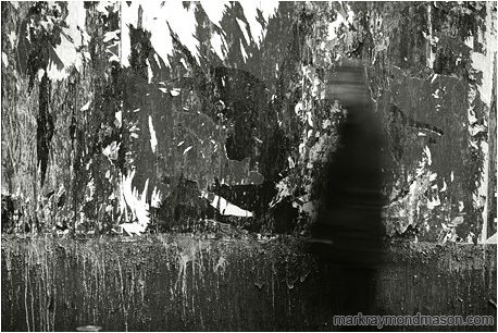 Abstract black and white photograph of a blurry figure walking past the chaotic shapes of advertisements that have been torn from a wall