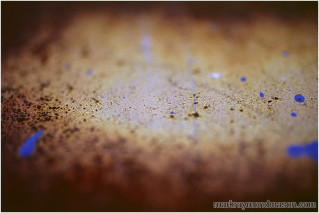 Abstract photograph of blue paint splatter seeming to hover above a blurry, rusted steel plate