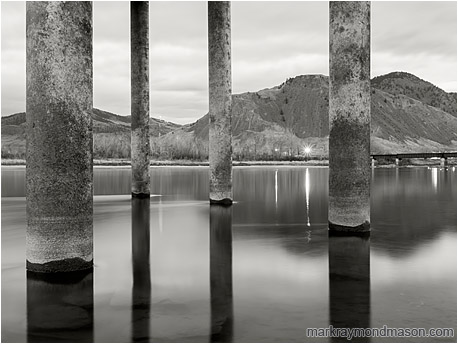 Fine art black and white photograph of large piles rising out of the frame from the smooth water of a full river