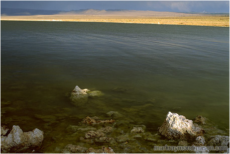 Fine art photograph of small volcanic rocks in a large lake, with stormclouds and sunshine in the distance