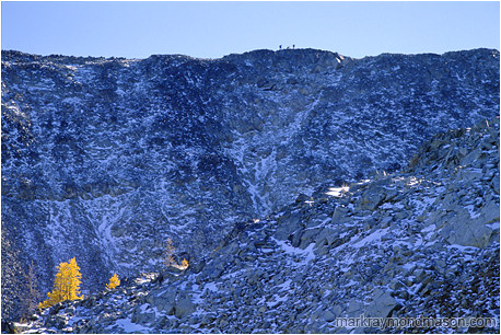 Lifestyle photo showing hikers high atop a rocky mountain ridge, with colourful larch trees below them