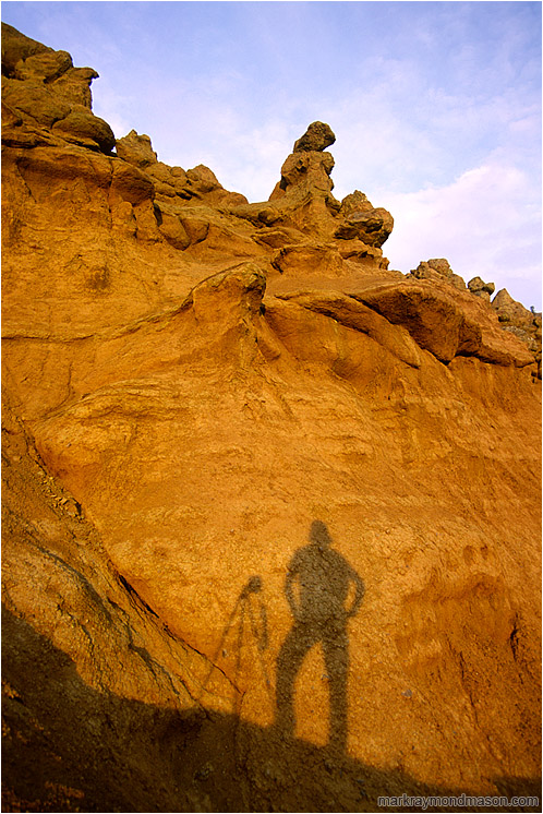 Self Portrait With Camera Shadow: Near Kamloops, BC, Canada (2002-00-00) - Landscape photograph of a silhouetted photographer and camera mounted tripod against a dramatic sculpted hoodoo background