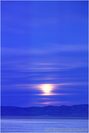 Landscape photo of a white moon rising through clouds over a dry desert lake