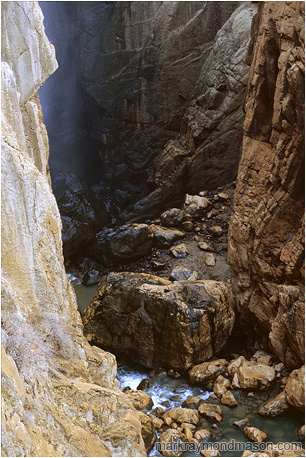 Fine art nature photograph of water and mist in a deep slot canyon, with steep cliffs of orange and black rock