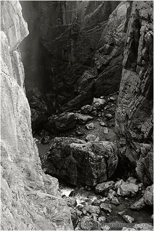 Fine art black and white photograph of a deep, steep-sided canyon, a misty waterfall, and a rushing river