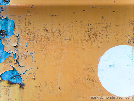 Abstract fine art photo of a burning white circle painted on scrap metal, the orange flaking to reveal a fine blue layer underneath