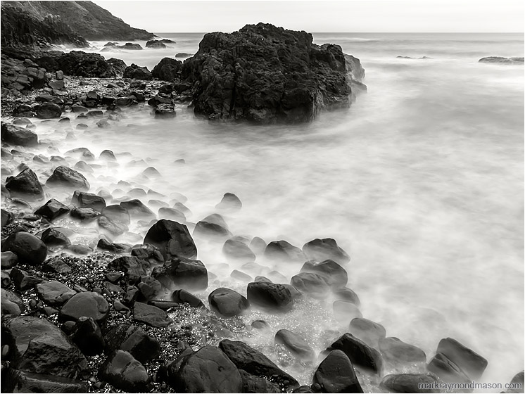 Worn Rocks, Crashing Waves: Near Stawberry Hill Park, OR, USA (2015-10-21) - Fine art black and white photograph of smokey waves swirling around a rocky knob and a bounder-strewn beach