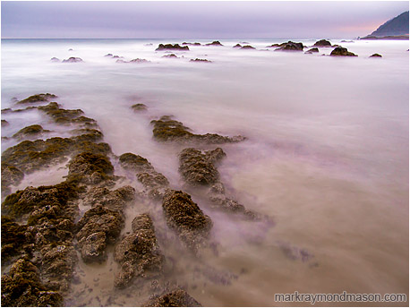 Fine art long-exposure photograph of barnacled rocks and clouds of sand as seawater washes over a tidepool