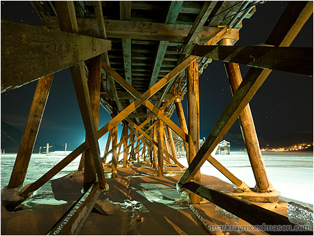 Fine art photograph showing giant beams on the underside of a wharf, frozen in ice, set against a night sky with faint stars
