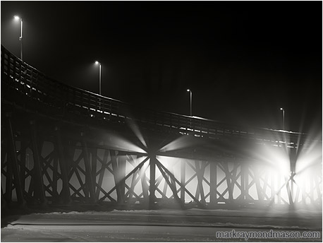 Fine art black and white photo showing light streaming through the crossed beams of an ice-locked wharf in the night