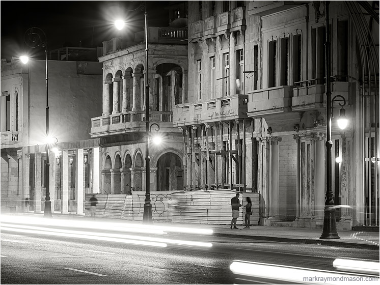 Buildings, Streaked Lights, Figures: Havana, Cuba (2017-02-13) - Fine art black and white photograph showing 2 figures beneath colonial architecture, through white streaked headlights