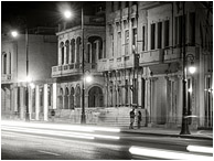 Buildings, Streaked Lights, Figures: Havana, Cuba (2017) - Fine art black and white photograph showing 2 figures beneath colonial architecture, through white streaked headlights