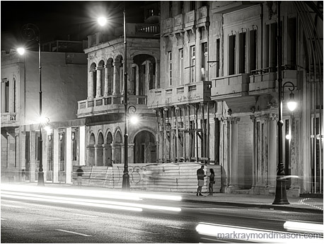 Fine art black and white photograph showing 2 figures beneath colonial architecture, through white streaked headlights