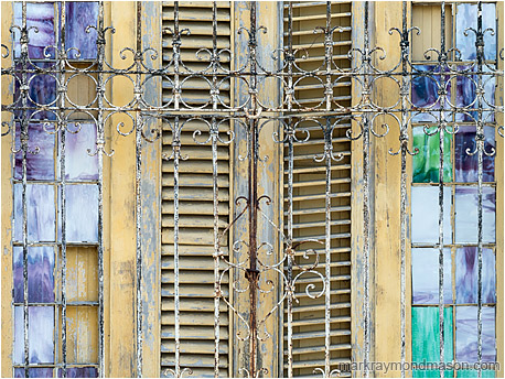 Abstract photography showing small stained glass windows and wooden shutters behind ancient wrought-iron bars
