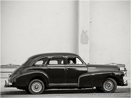 Fine art black and white photograph showing silhouetted lovers in a battered 1950s model car, parked beside a plain concrete wall