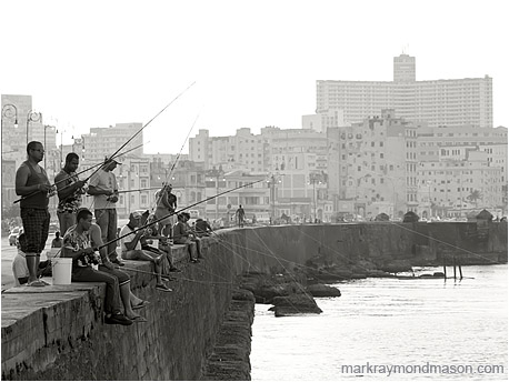 Black and white landscape photograph showing men fishing from the Malecon, with the ancient buildings of Havana in the background