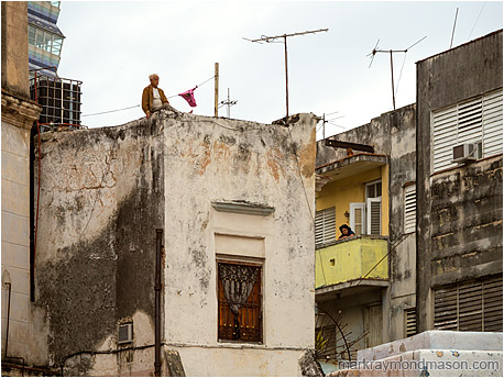 Fine art photograph showing a man perched unsettlingly on top of a concrete building and a woman looking into the street from a mid-level apartment