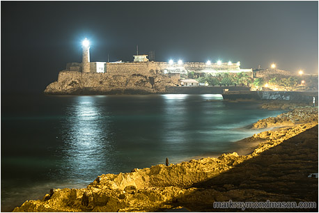 Fine art photograph of a figure on the rocks by the sea, with floodlights on a fortress and a lighthouse in the background