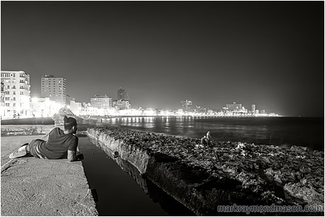 Fine art black and white photograph of a man resting on the seawall in Havana, taking in the city lights and a party fishing by lamplight