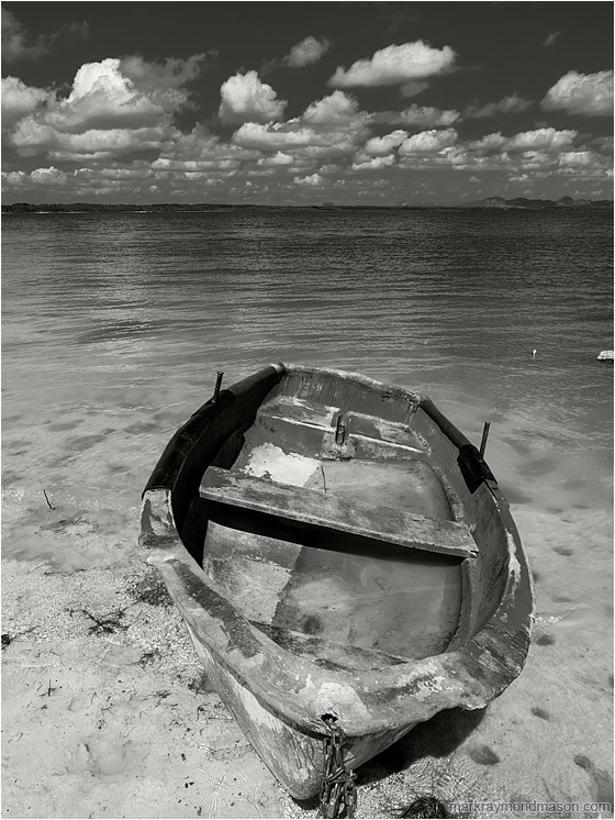 Battered Boat, Rolling Clouds: Near Vinales, Cuba (2017-02-23) - Fine art black and white photograph showing a flooded boat, cocked sideways, sitting abandoned on the silty shores of a shallow lake