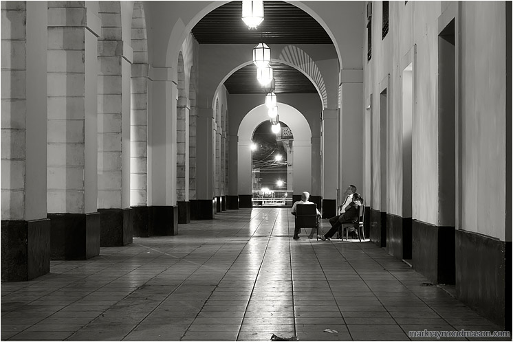 Arches, Resting Guards: Matanzas, Cuba (2017-02-26) - Fine art black and white photograph showing several bank guards lounging in chairs in an empty breezeway