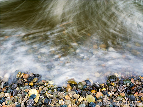 Fine art photograph showing intertwined, blurry waves moving over round multi-coloured beach rocks