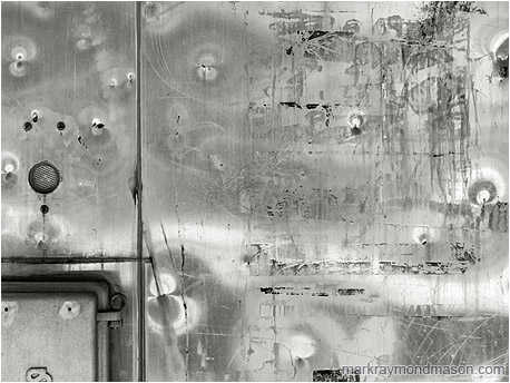 Fine art abstract black and white photo showing a closeup of a dented metal train kiosk, reflected light forming patterns in the surface